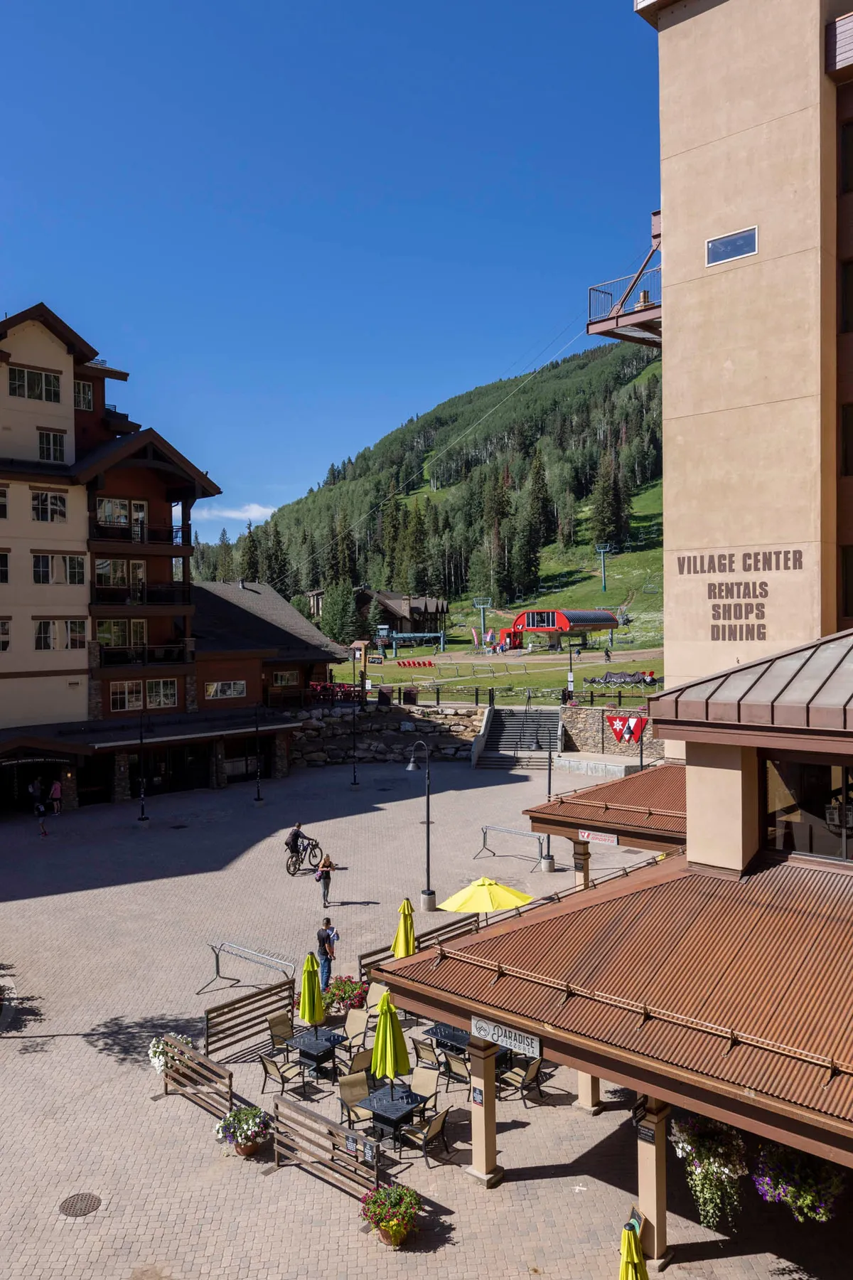 The image captures the heart of Village Plaza, featuring an inviting outdoor seating area dotted with vibrant yellow umbrellas. Surrounding the plaza are shops and a central building, bustling with people enjoying shopping, dining, and biking. The backdrop of scenic mountains reinforces the sense of a lively, well-situated community.