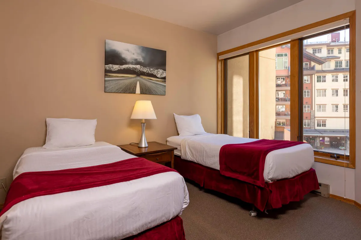 This image features two twin beds with red bed skirts, a table lamp, a large window offering a city view, and mountain landscape artwork on the wall. The morning light fills the room, emphasizing the cozy and comfortable ambiance.
