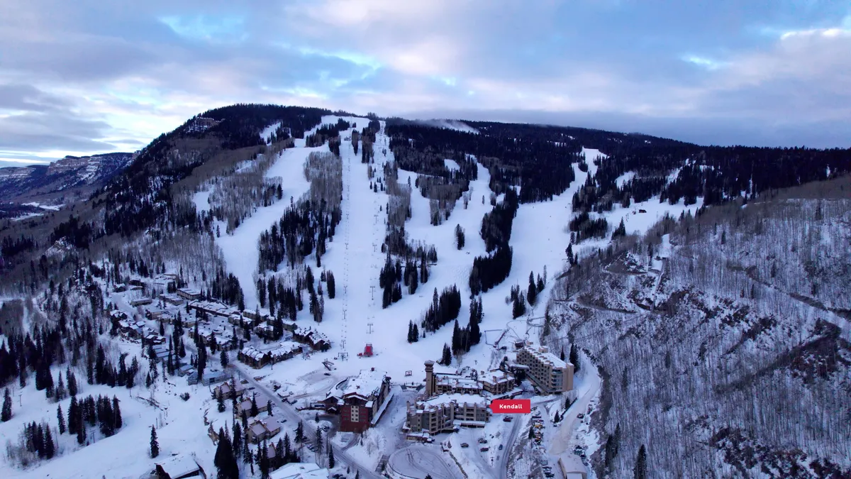 The image depicts the exterior of residential buildings set against a backdrop of ski slopes and pine trees, evoking an adventurous, active mountain resort atmosphere. The snow-covered landscape hints at the proximity to Purgatory Resort’s activities and village amenities.