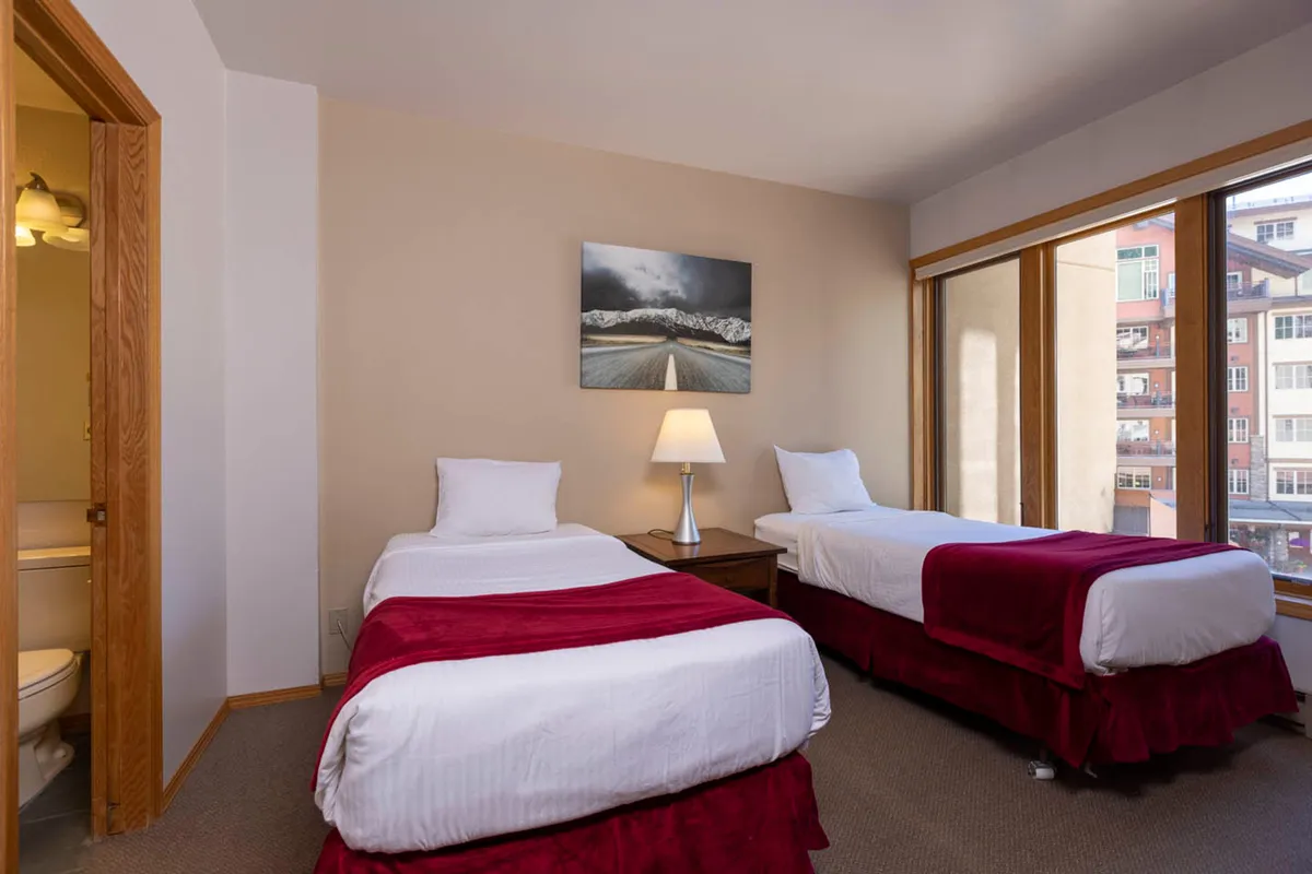 A cozy bedroom with twin beds, cheerful wall art, a reading lamp on the nightstand, and large windows that fill the space with natural light—perfect for unwinding and enjoying the mountain surroundings.