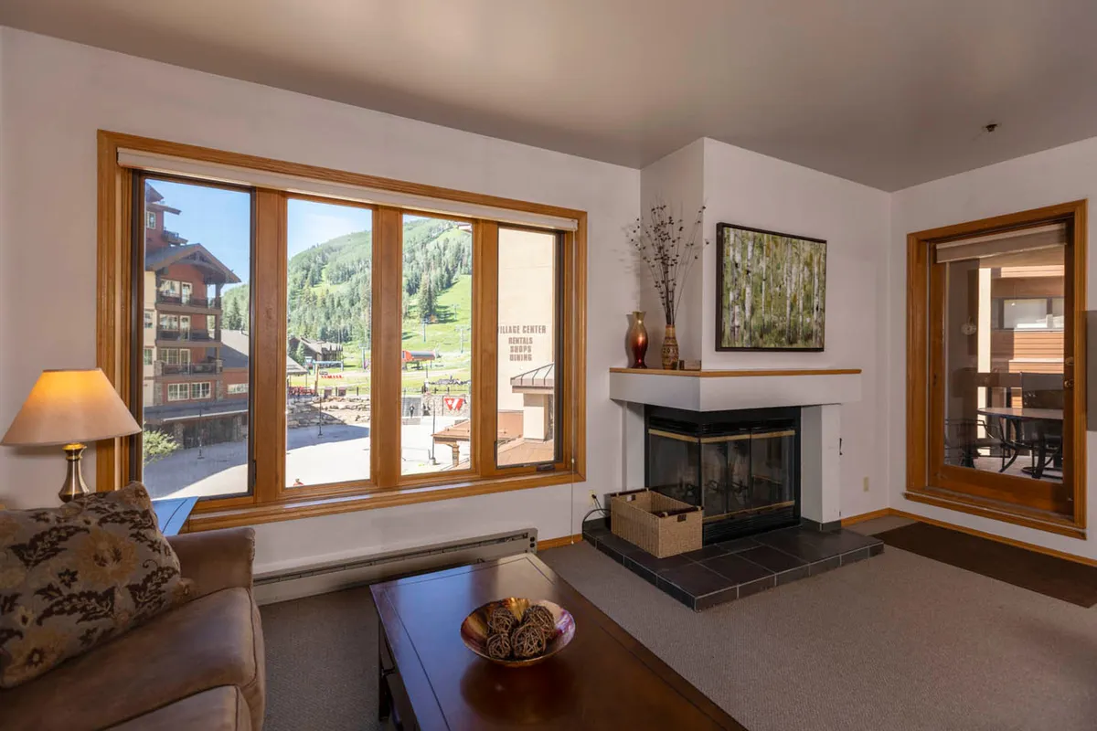 A spacious living room with a wood-burning fireplace as its centerpiece, flanked by large windows offering stunning mountain views. A comfortable sofa and coffee table create a welcoming area perfect for relaxing, socializing, or enjoying the warmth of the fire after a day of adventure.