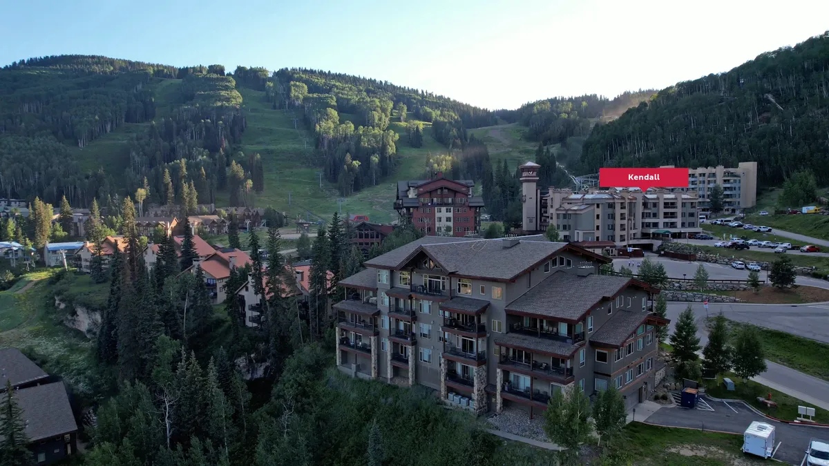 Overlooking residential buildings set against a peaceful mountain backdrop, the image highlights the close relationship between the property, green spaces, and the parking area in front. The tranquil vibe is balanced by visible community activities in the plaza.