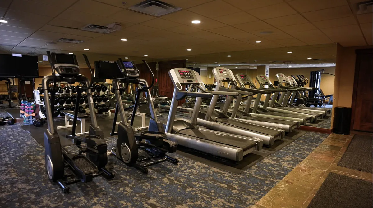 The image presents a modern fitness center interior with treadmills, ellipticals, weights, and mirrors. The space is bright and motivational, designed to support both individual workouts and group fitness activities.