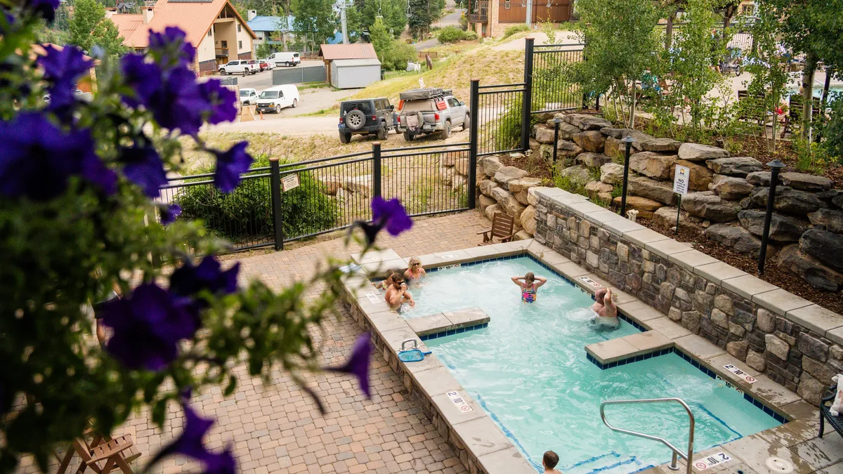 This image showcases a vibrant outdoor swimming pool area surrounded by flowers and a rock wall, with people enjoying the water and sunbathing. The lively setting emphasizes leisure and socializing in a picturesque mountain resort environment.