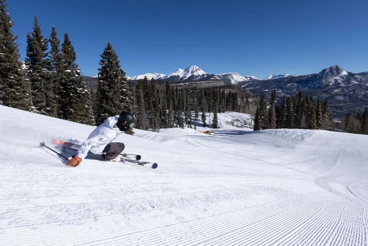 A skier glides down a snow-covered slope framed by majestic mountains and trees, with the crisp winter air and brilliant sunlight capturing the vibrant spirit of the area. The expansive outdoors hints at exciting activities and breathtaking landscapes awaiting guests just steps from the resort.