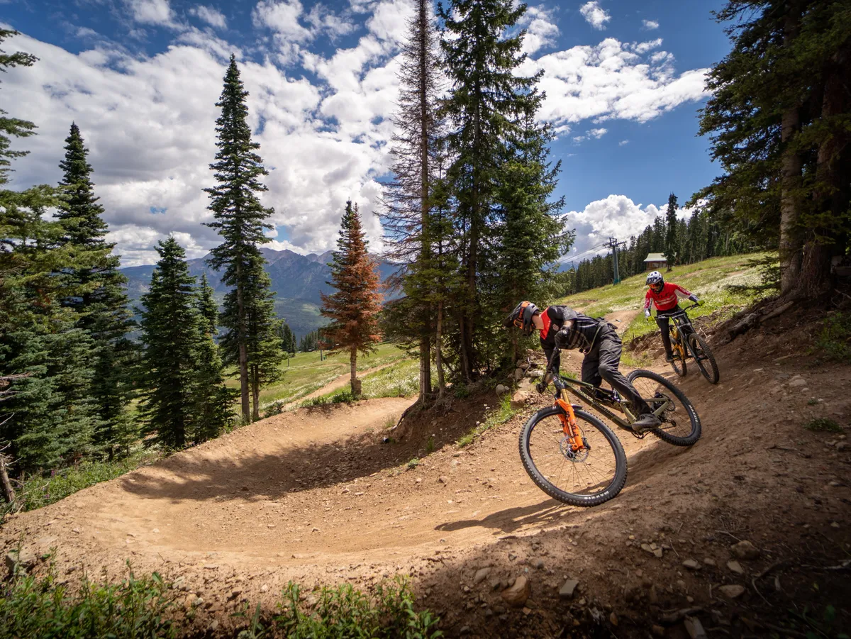 An energetic scene of mountain bikers racing along a curved dirt path framed by lush trees and dramatic mountain backdrops. The action-filled composition highlights summer adventures and the natural beauty surrounding the resort.