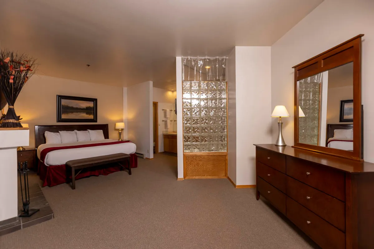 This image showcases a serene bedroom featuring a comfortable bed with a decorative throw, a wooden dresser, a glass block wall, and a lamp on the bedside table. The setting is bathed in soft lighting, creating a peaceful and inviting space perfect for relaxation.